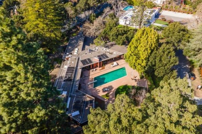 An aerial view of an Atherton Eichler: a low dark-roofed house with solar panels arranged in wings around a swimming pool and terrace, surrounded by mature trees.