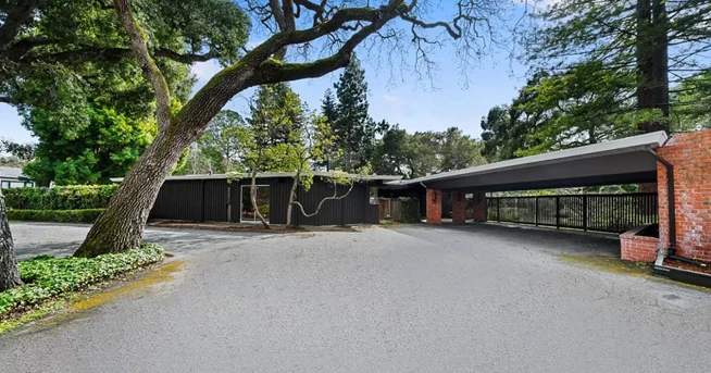 A dark-stained single-storey house behind a fence, with an open carport on brick piers under a flat roof, beneath a leaning oak.