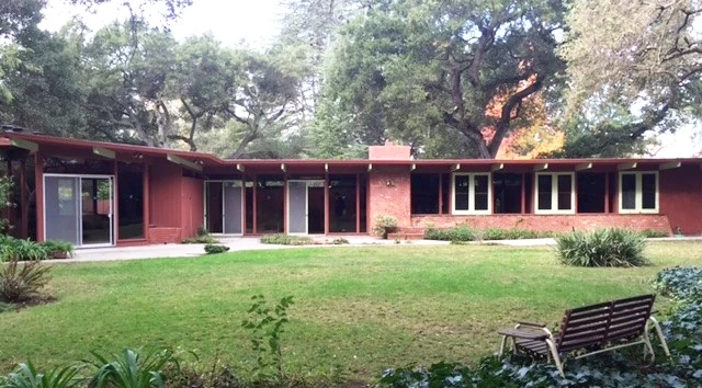 A single-storey Eichler stained deep red, with a run of windows and sliding doors under a flat roof carried on exposed posts, seen across a lawn with a bench.