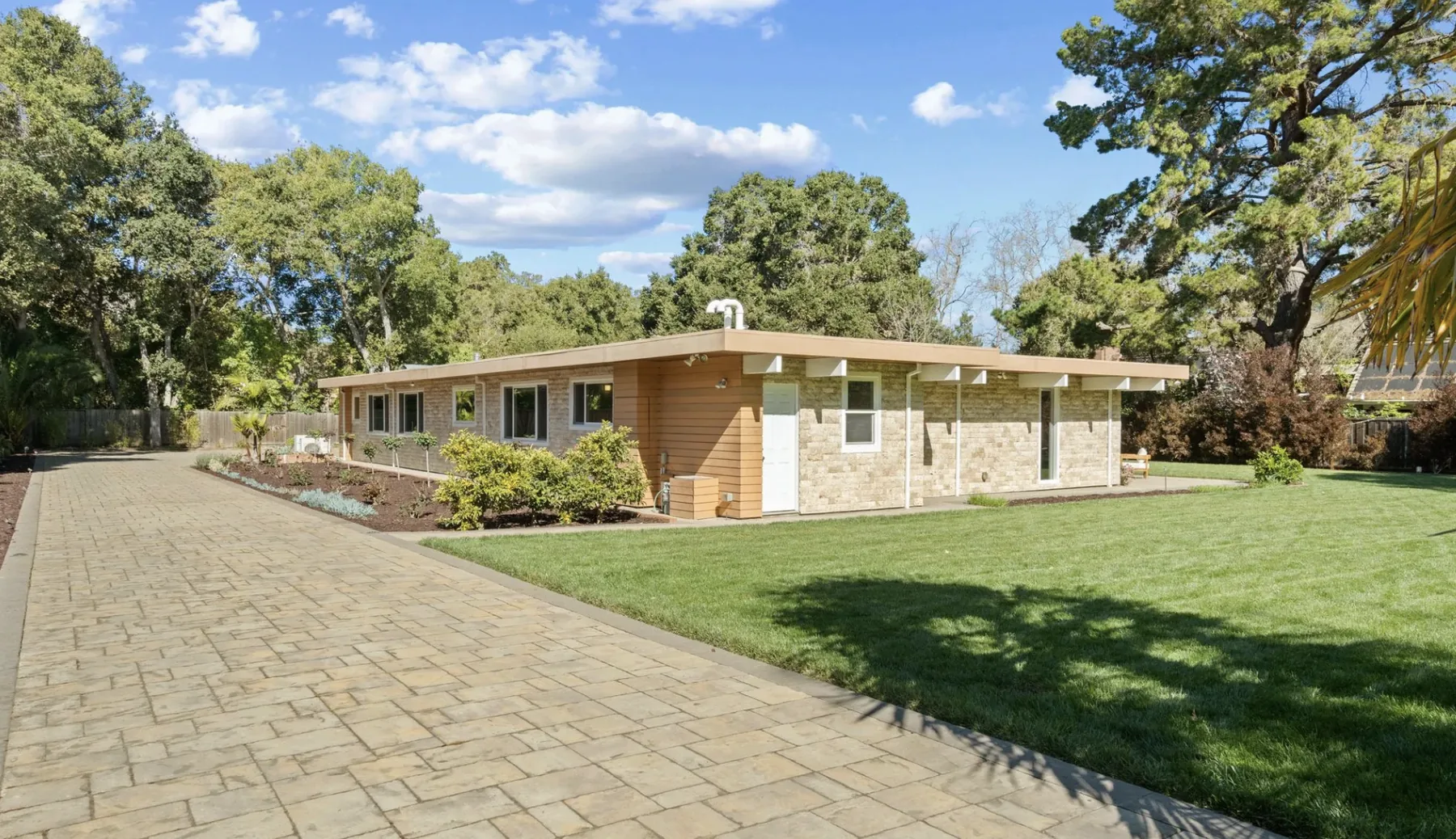 The entrance elevation of an Atherton Eichler from a paved motor court: a long low wall of stone and horizontal boarding under a flat overhang, with small high windows and a plain white door.