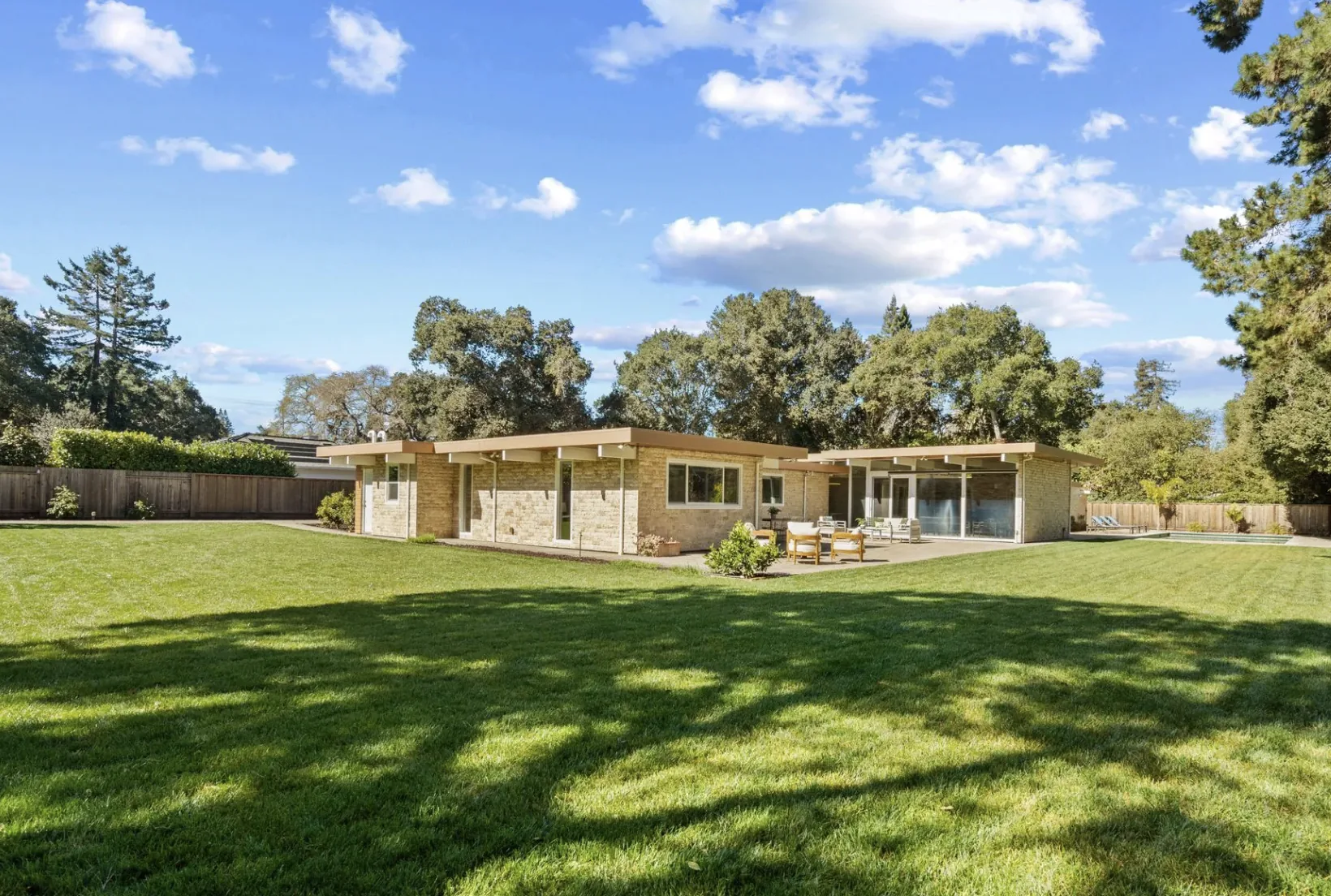 An Atherton Eichler seen from across a large lawn: a low house with wide flat roof overhangs, stone-clad walls and glass sliders, bounded by a wooden fence, with a pool at the right.
