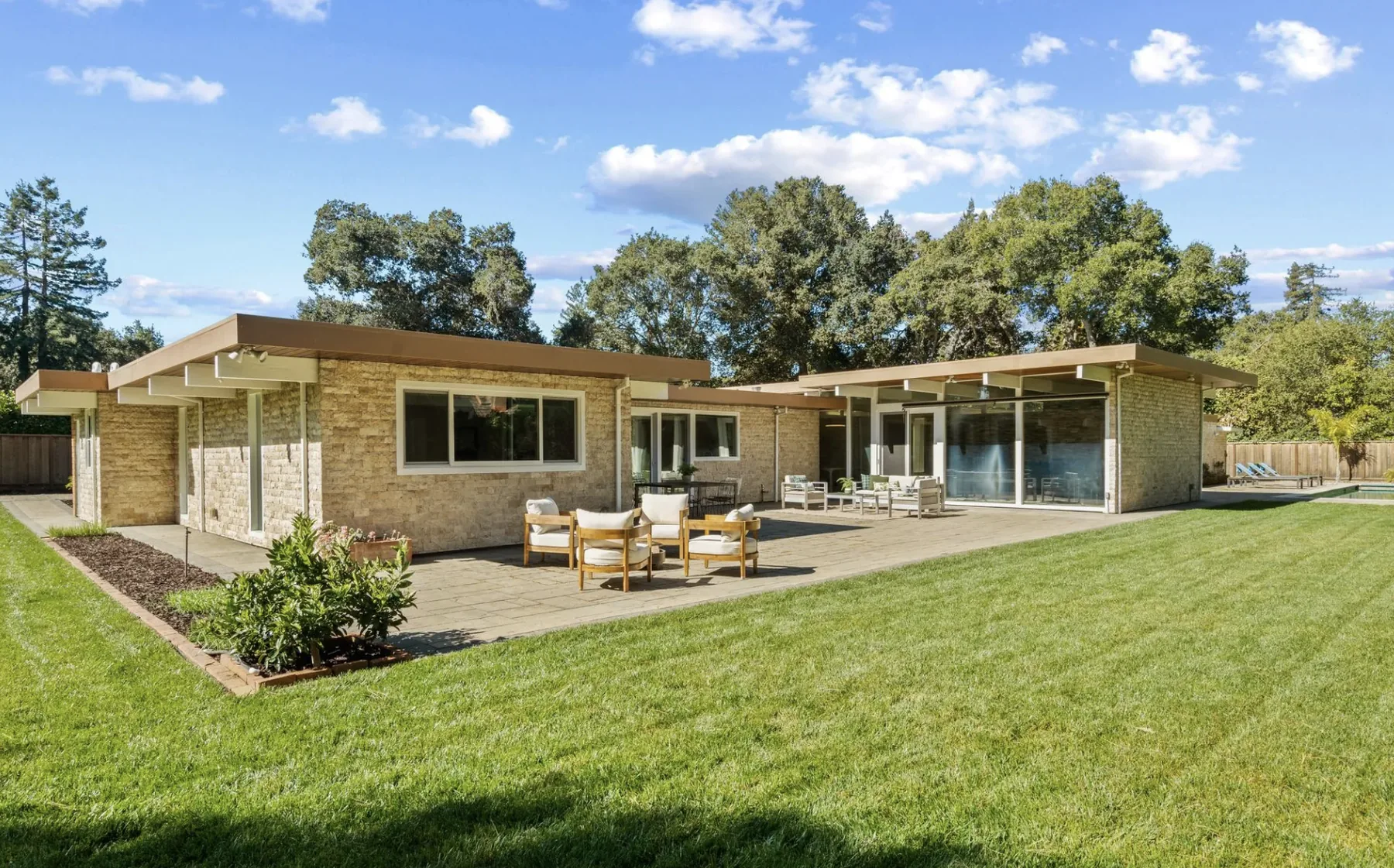 An Atherton Eichler from its garden: a long single-storey house with wide flat overhangs on slender posts, stone-clad walls, a run of glass sliding doors onto a paved terrace with outdoor furniture, and a lawn and pool beyond.