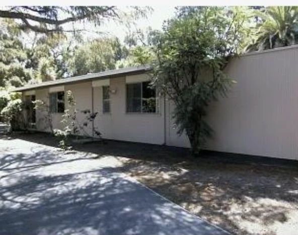 A low white-painted house at the end of a drive, with a flat roof, plain windows and a white fence, screened by trees.