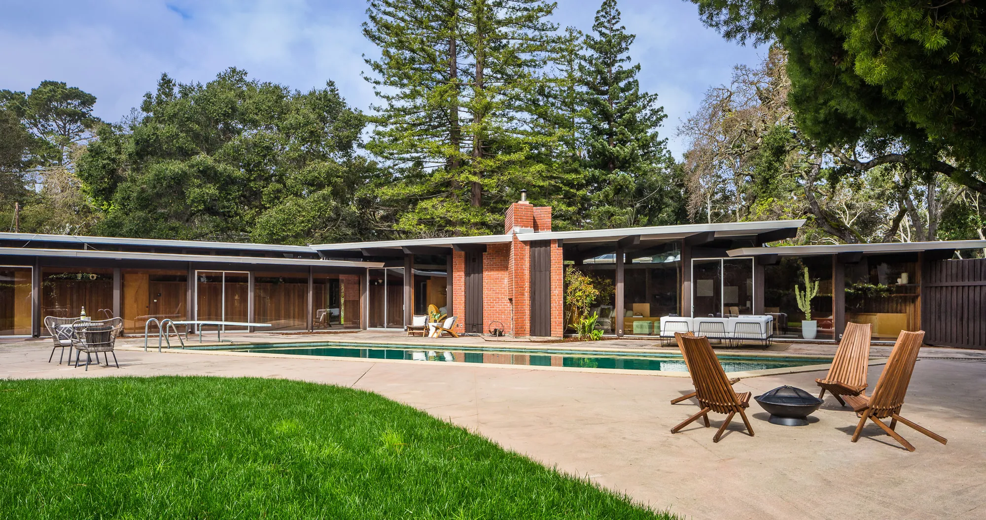 19 Irving Avenue by day: a long single-storey house of glass walls under low white-edged roofs, a brick chimney at its centre, a swimming pool and terrace in front and lawn in the foreground.