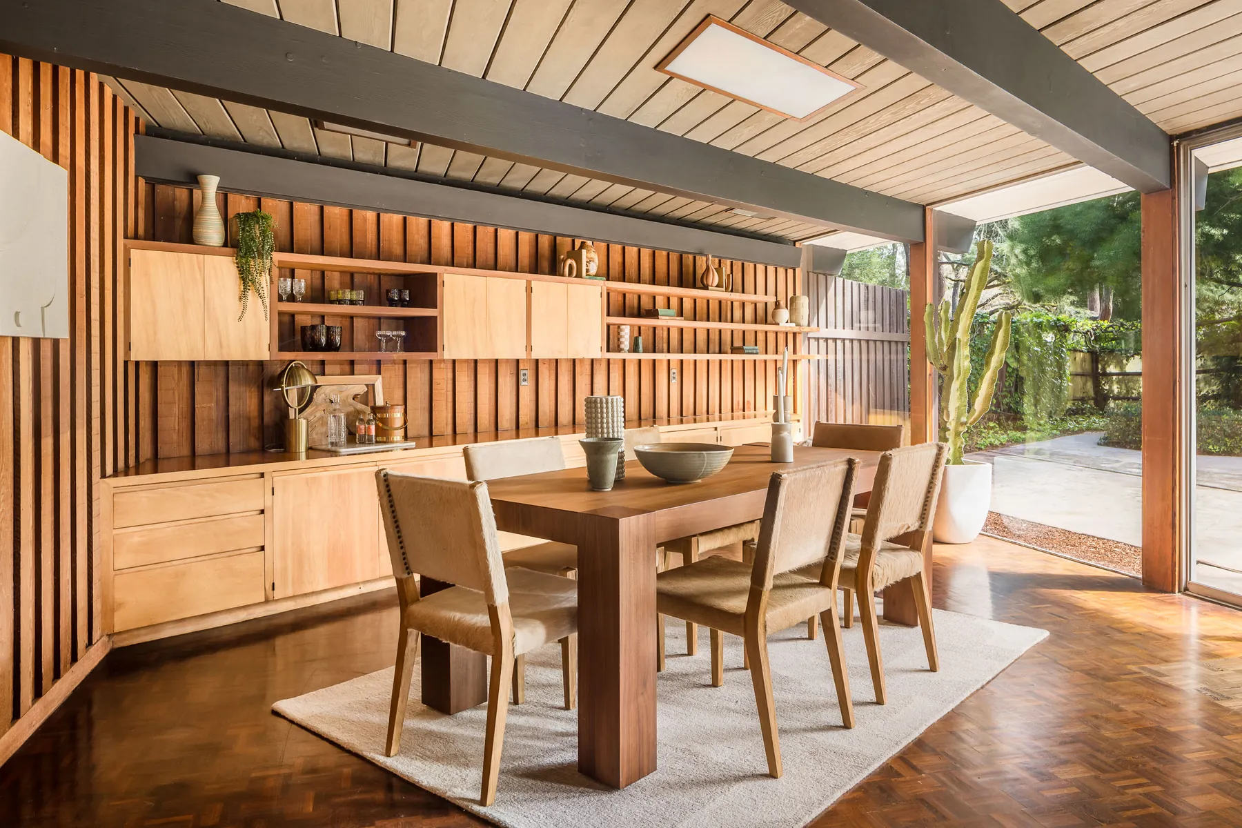 Dining room with a long built-in wall of blond wood cabinets and open shelves set into vertical redwood panelling, a wooden table and chairs, and a full-height glass wall to the garden.
