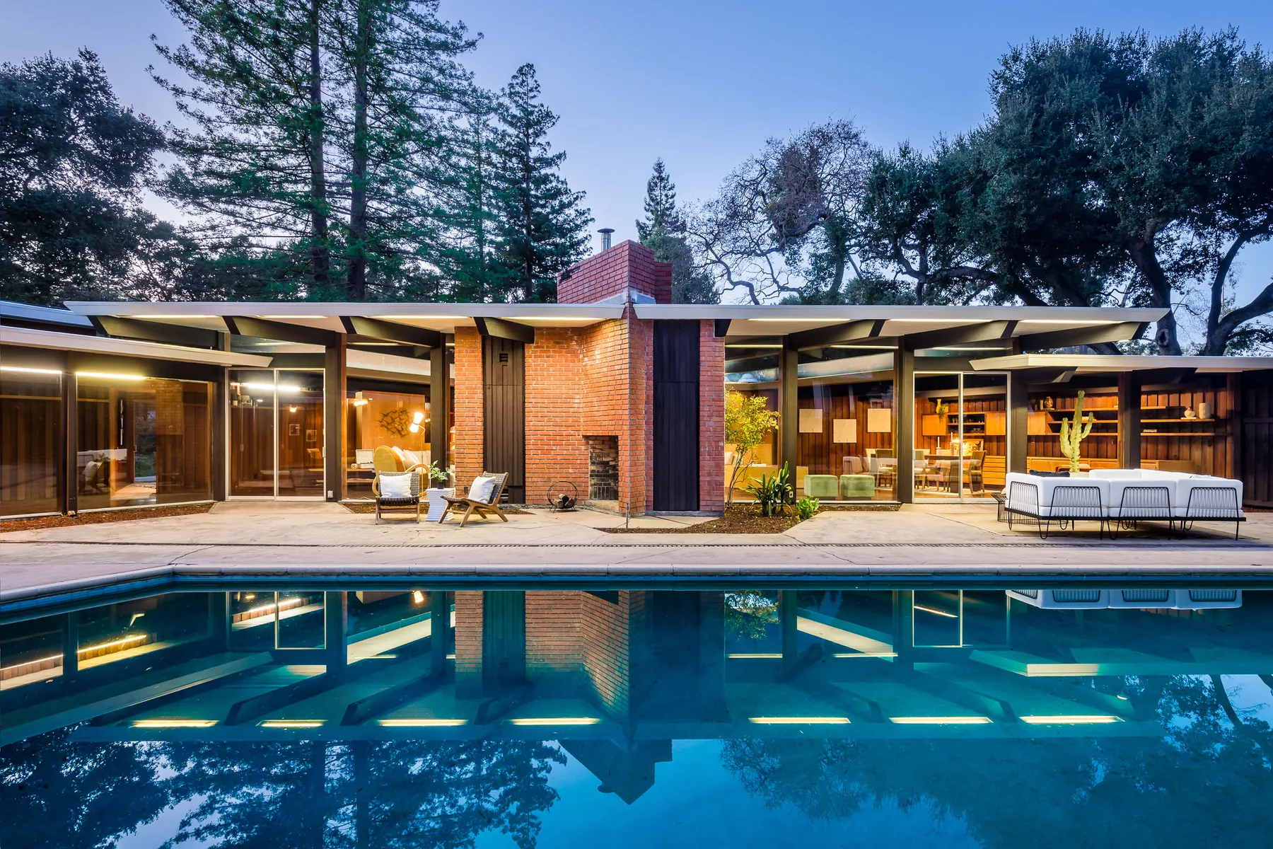 The garden front at dusk reflected in the swimming pool, lights on inside the glass walls and the brick chimney at the centre.