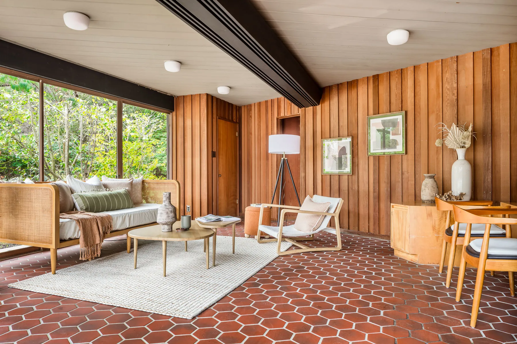 A family room with a red hexagonal tile floor, vertical redwood panelling, a large window onto trees, a cane daybed and a white rug.