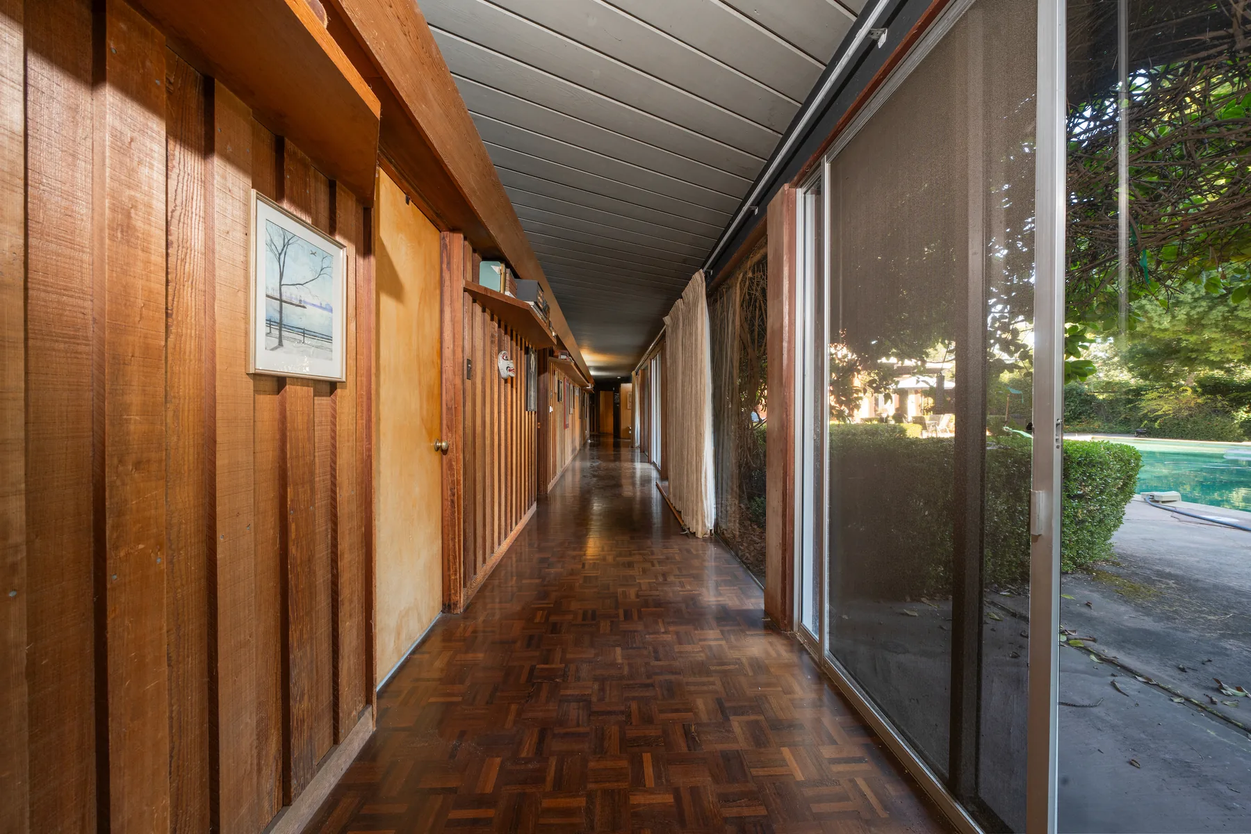 A long interior corridor with a parquet floor, vertical redwood panelling and a built-in shelf on the left, and a wall of sliding glass on the right looking onto the pool.