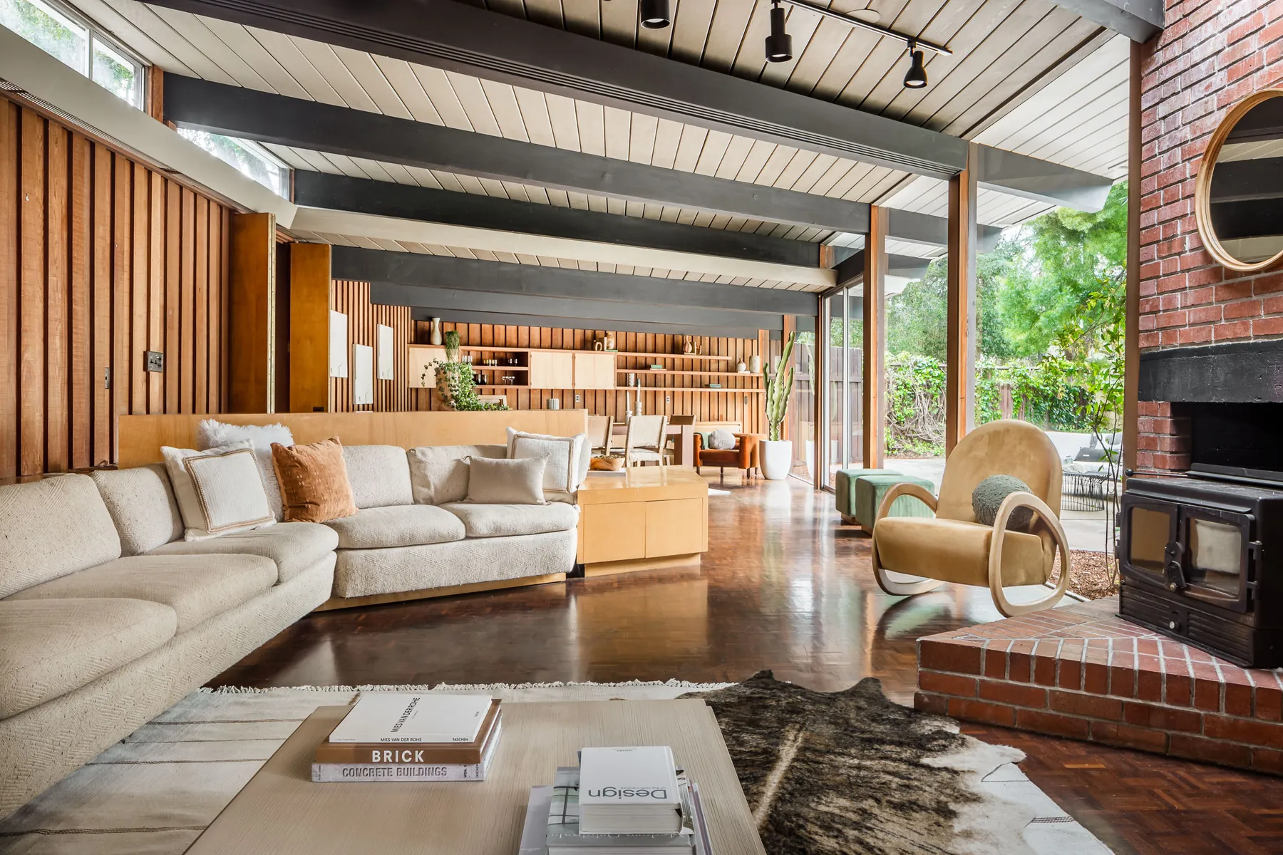 The living room at 19 Irving Avenue: exposed dark beams under a pale board ceiling, clerestory windows over redwood panelling, parquet floor, and a full-height glass wall onto the garden beside a brick hearth.
