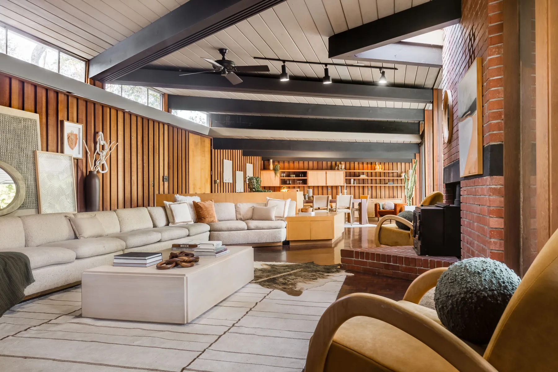 The living room: a long sectional sofa under dark exposed beams and a pale board ceiling, vertical redwood panelling with high windows above, and a brick fireplace wall on the right.