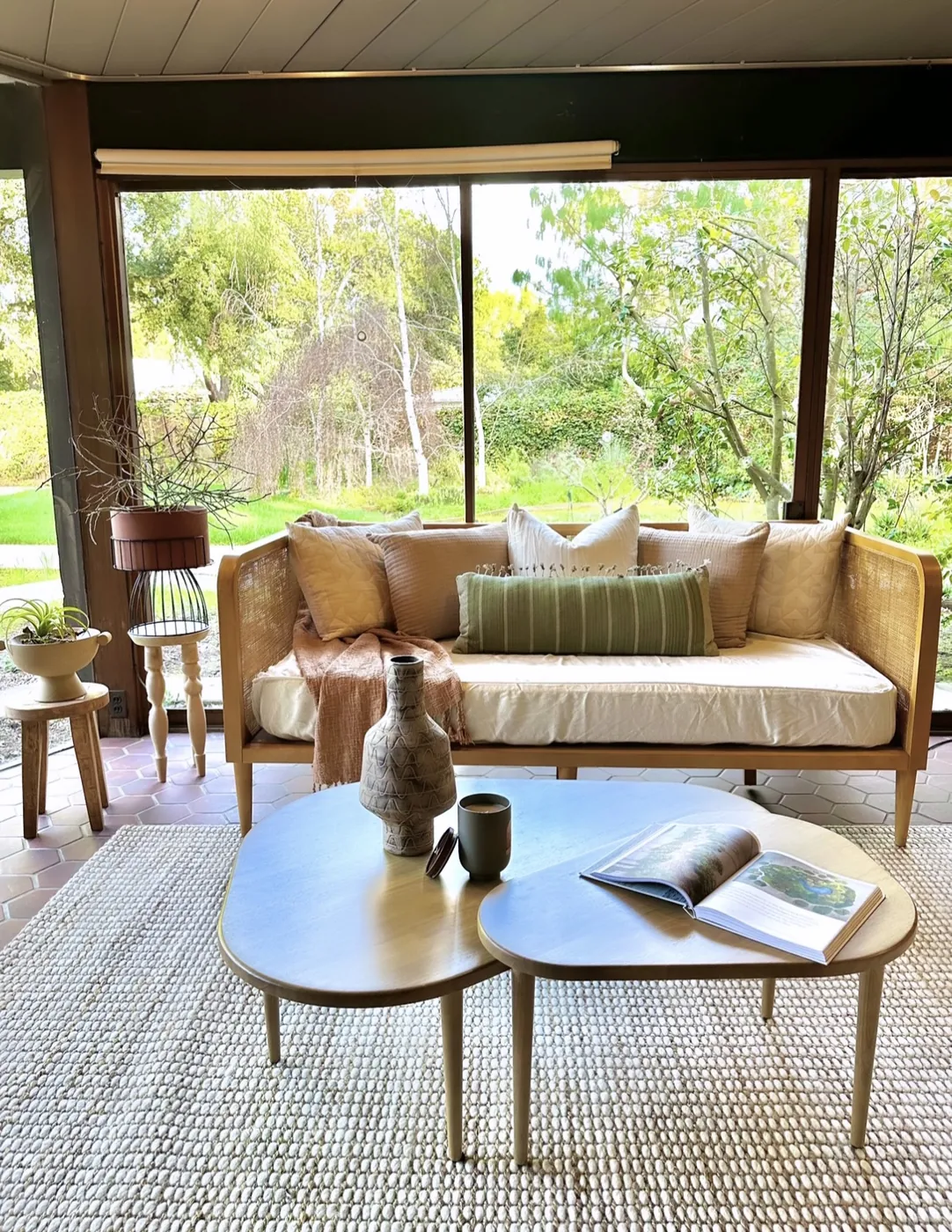 A cane daybed on a red hexagonal tile floor in front of a full-height glass wall looking onto a lawn and trees, with a low kidney-shaped table in front.