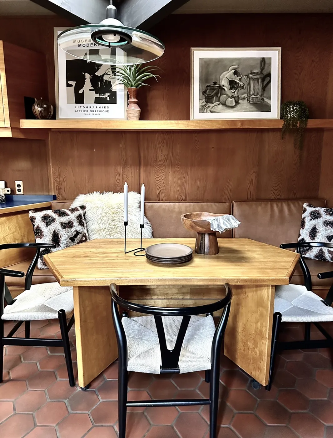 A hexagonal wooden dining table and black chairs at a built-in leather banquette, against a plywood wall with a long shelf above it, on a red hexagonal tile floor.