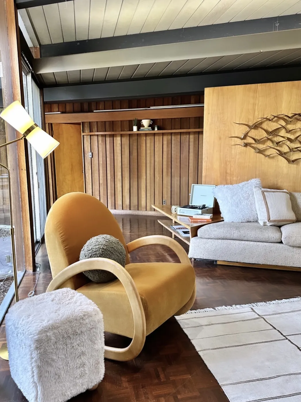A lounge chair and sofa on a parquet floor against vertical redwood panelling, with a large plywood partition beyond carrying a metal bird sculpture.