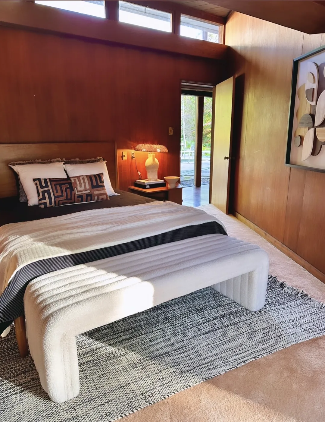 A bedroom panelled in warm wood with a clerestory window band at ceiling height, a bed with a wooden headboard, and an open door onto a paved terrace.